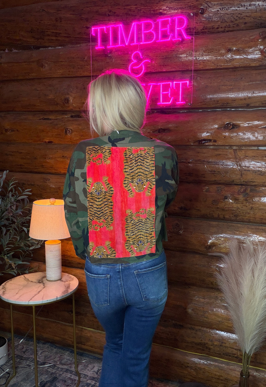 Person wearing a vintage military camouflage jacket with a pink pattern on the back, standing in front of a wooden wall with 'Timber & Velvet' neon sign.