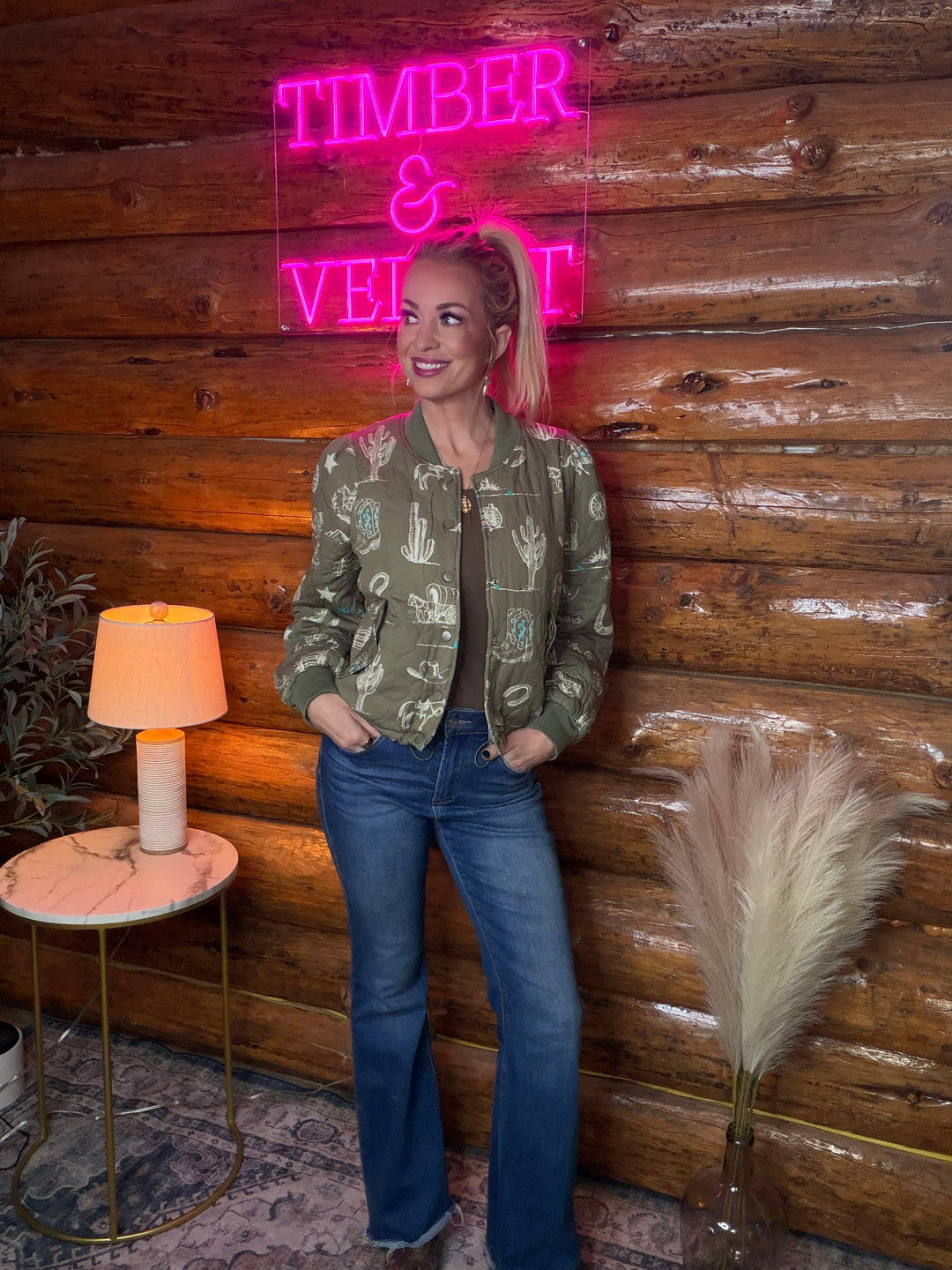 Person standing in front of a wooden wall  wearing a western theme quilted jacket with a neon sign reading 'TIMBER & VEGETABLE'.