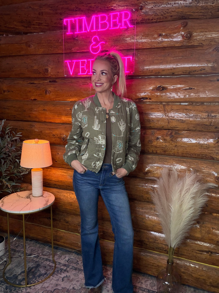 Person standing in front of a wooden wall  wearing a western theme quilted jacket with a neon sign reading 'TIMBER & VEGETABLE'.