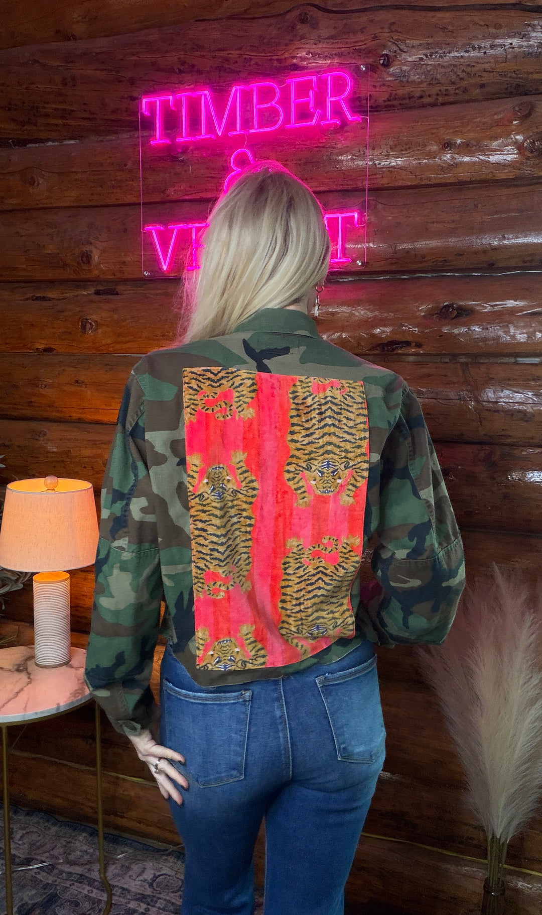 Person wearing a vintage military camouflage jacket with a colorful back pink panel with tigers in front of a wooden wall with a neon sign.