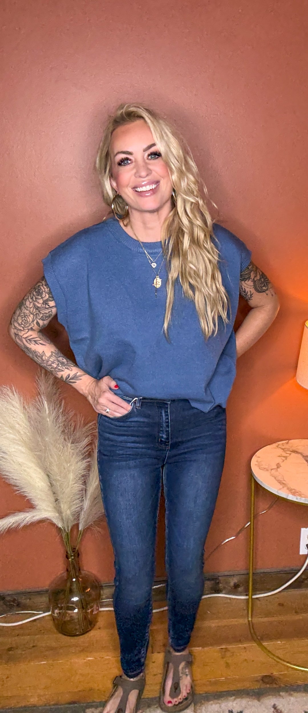 Woman in a blue top and jeans standing against a warm-toned wall.