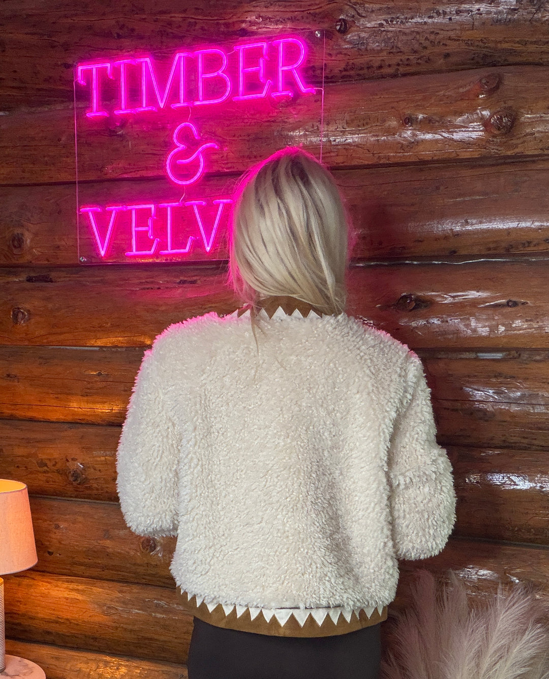 Person wearing a textured western themed white jacket in front of a wooden wall with a neon sign reading 'TIMBER & VELVET'.