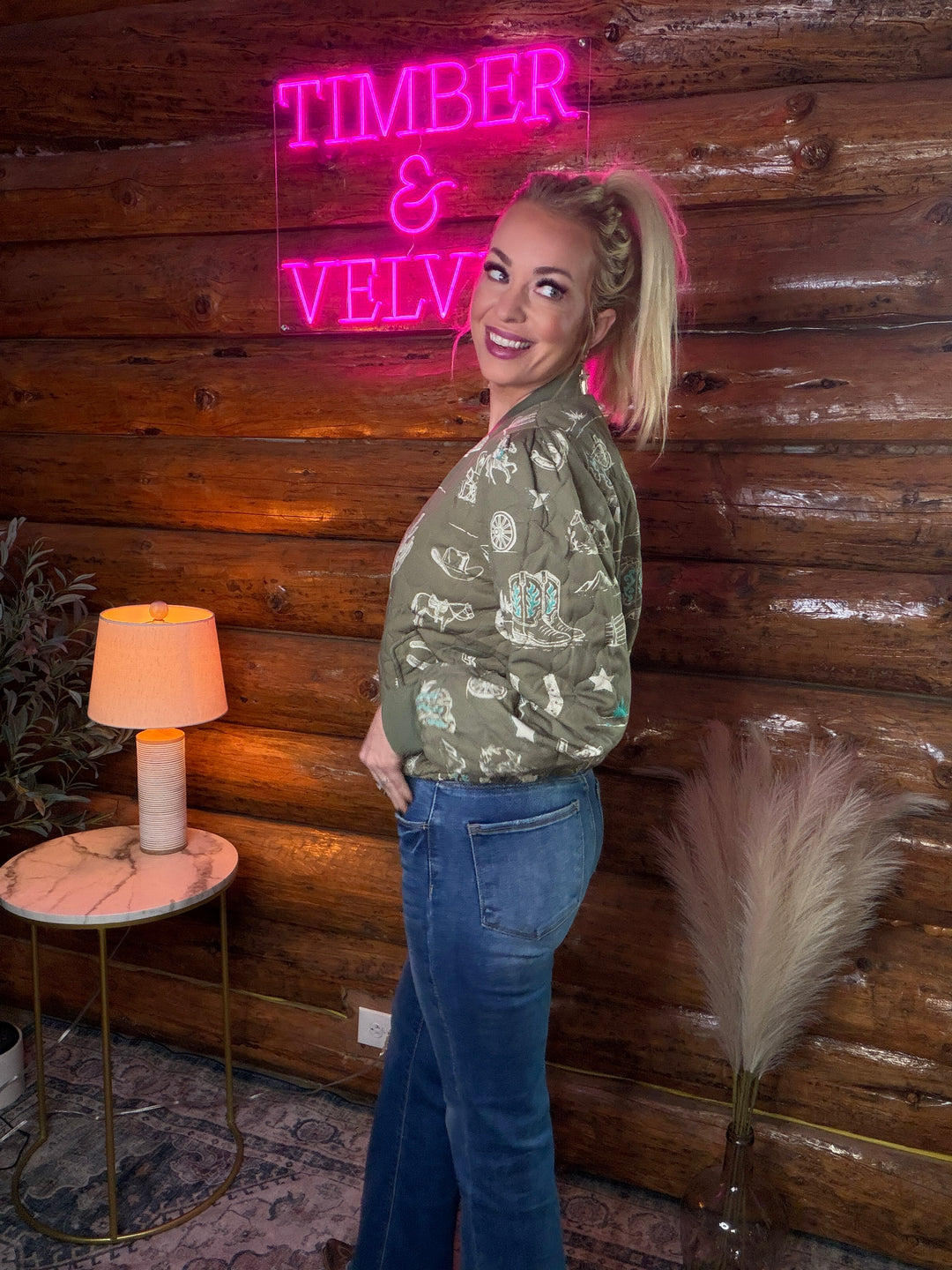 Woman wearing a western themed jacket standing in front of a wooden wall with a neon sign reading 'TIMBER & VELVET'.
