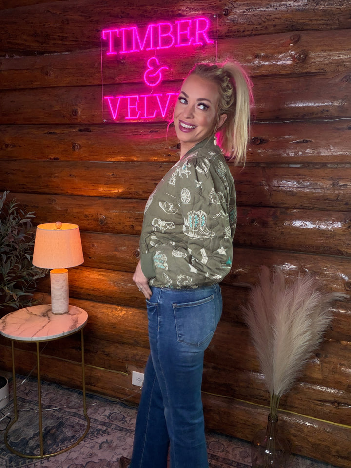 Woman wearing a western themed jacket standing in front of a wooden wall with a neon sign reading 'TIMBER & VELVET'.