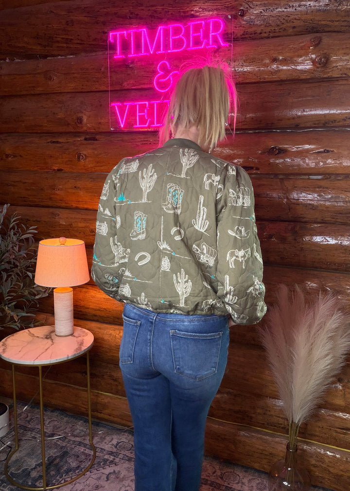 Person wearing a western themed jacket standing in front of a wooden wall with a neon sign reading 'TIMBER & VELVET’