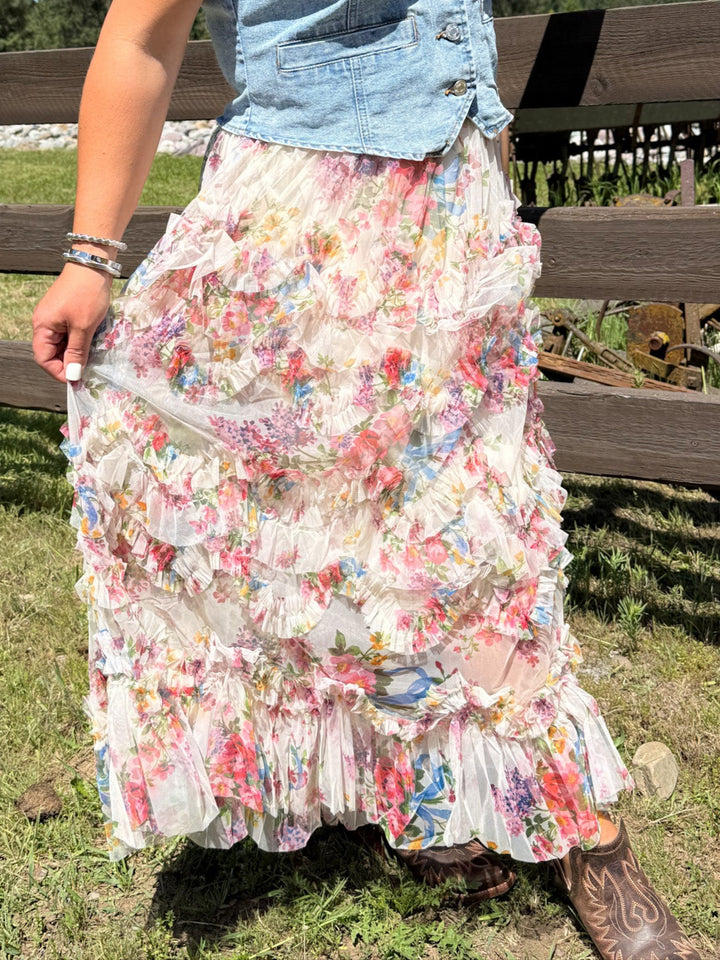 Model wearing a cascading ruffled tiers and a delicate blush-toned floral print skirt with a denim vest and boots.