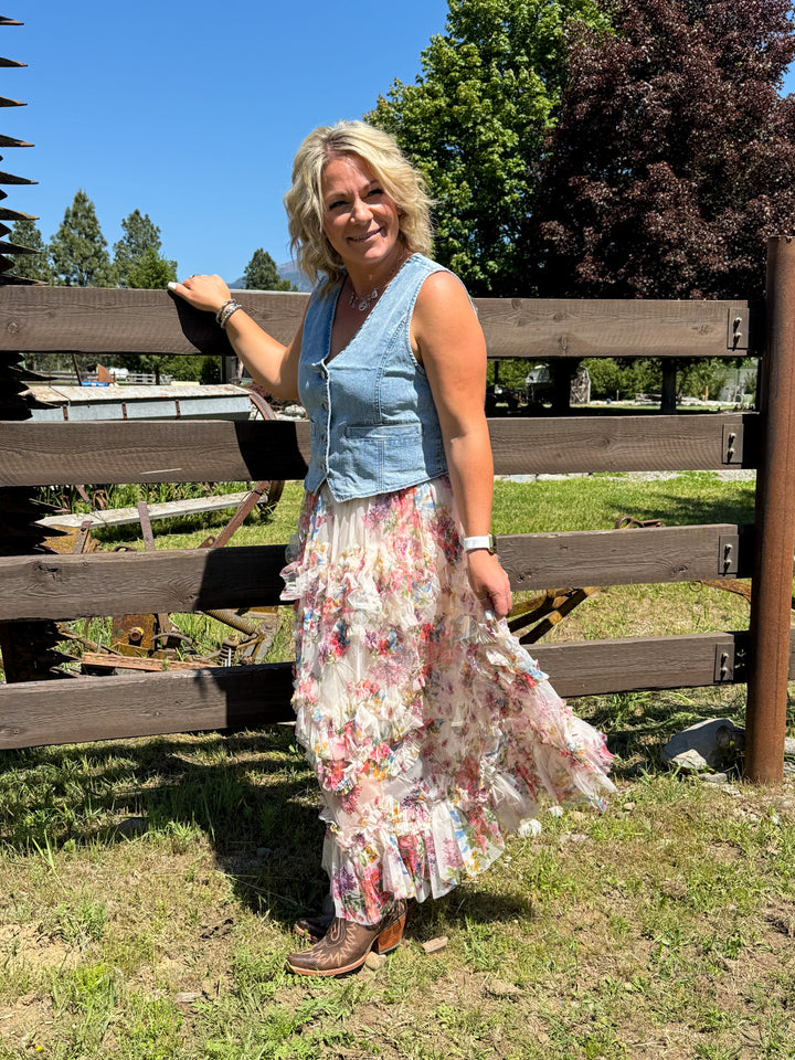 Model wearing a cascading ruffled tiers and a delicate blush-toned floral print skirt with a denim vest and boots.