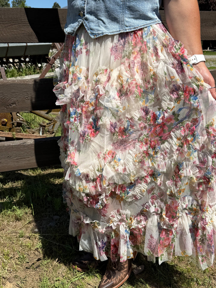 Model wearing a cascading ruffled tiers and a delicate blush-toned floral print skirt with a denim vest and boots.