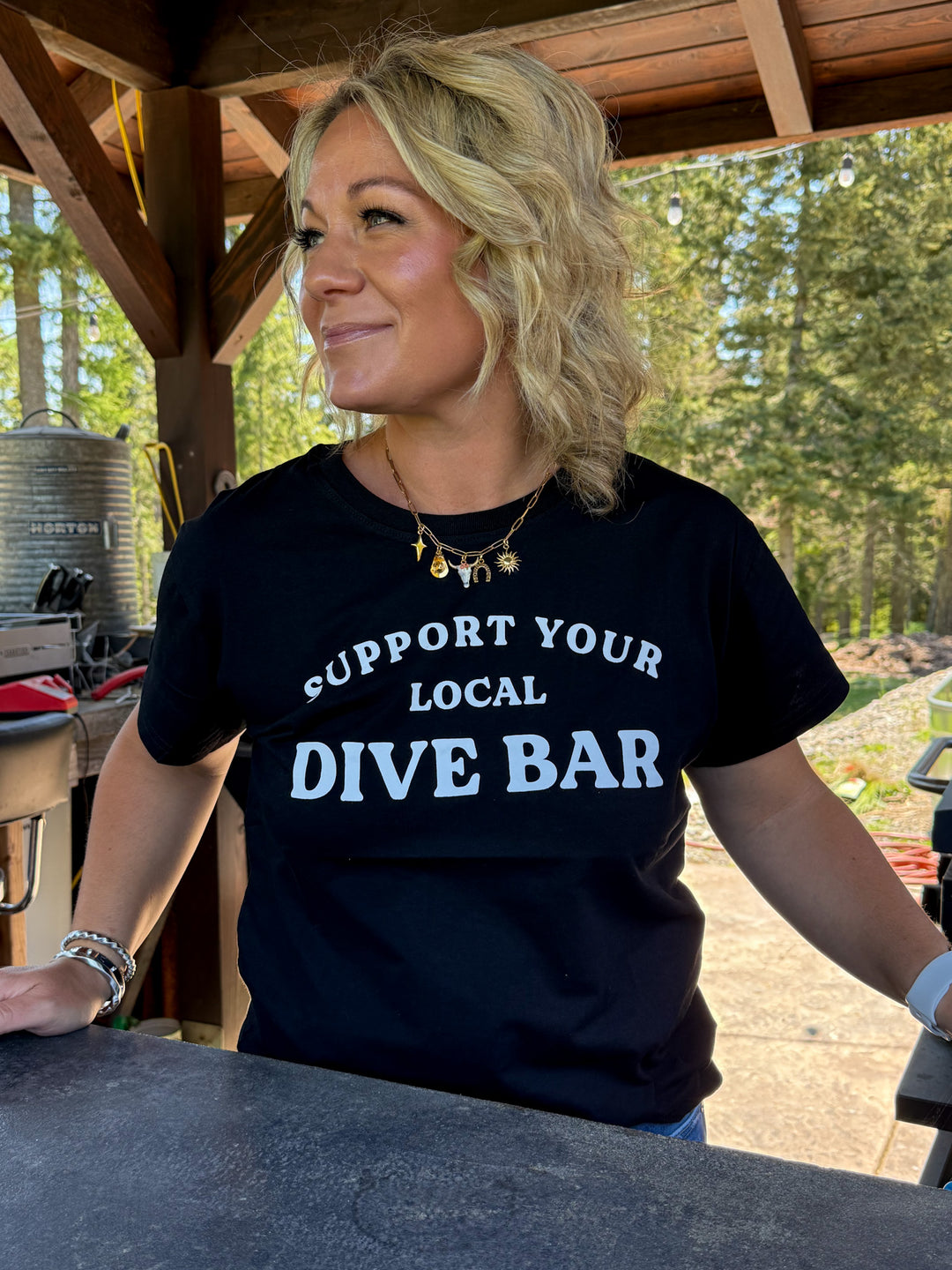 Model wearing black tee shirt with 'support your local dive bar' in white lettering across the front. Also wearing a western charm necklace in gold. 
