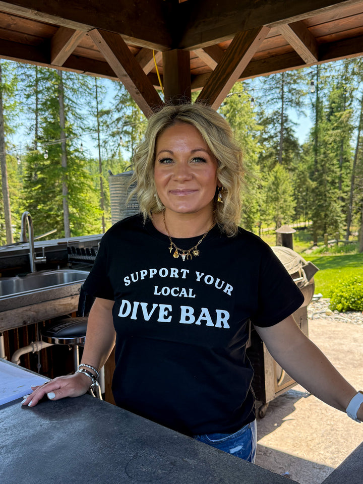 Model wearing black tee shirt with 'support your local dive bar' in white lettering across the front. Also wearing a western charm necklace in gold. 