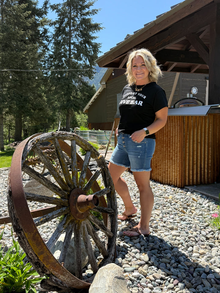 Model wearing black tee shirt with 'support your local dive bar' in white lettering across the front. Also wearing a western charm necklace in gold. 