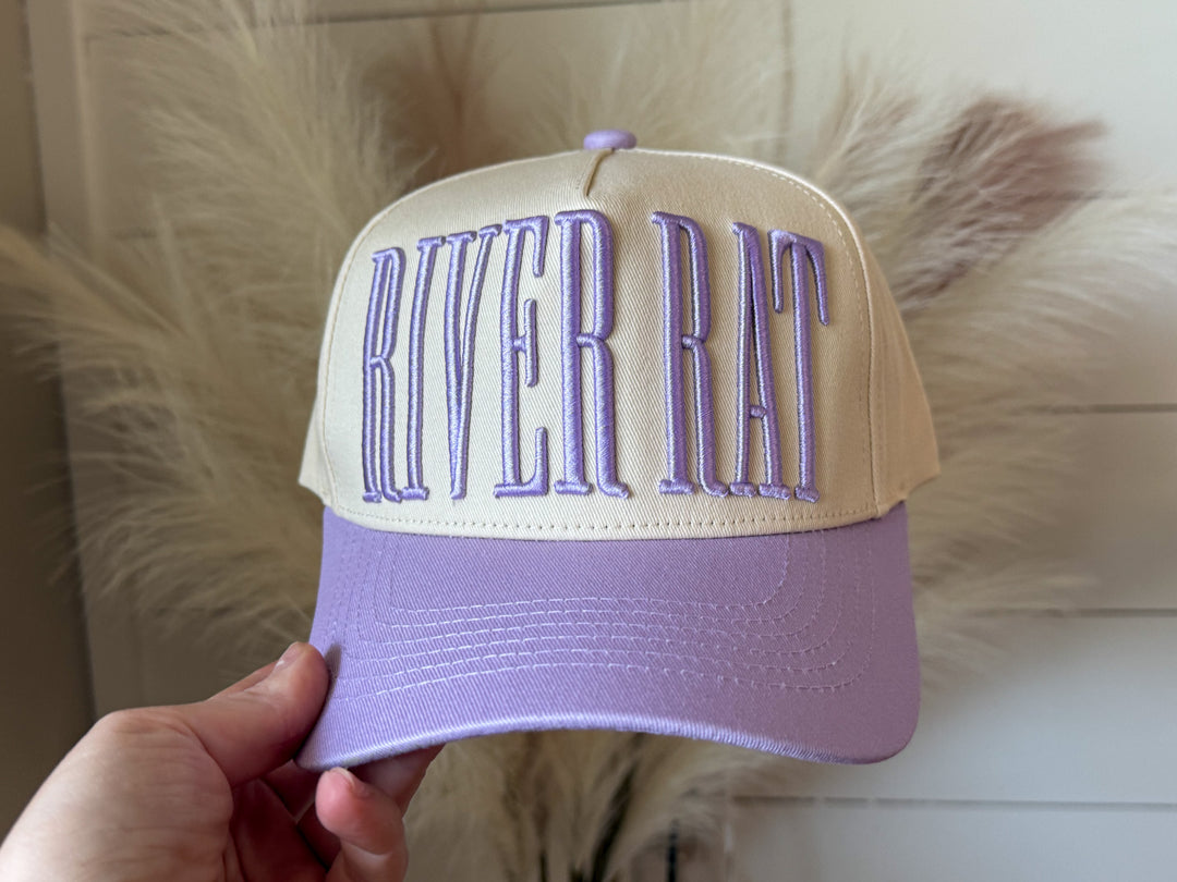 Lavender and cream colored trucker hat that has embroidered RIVER RAT across the front
