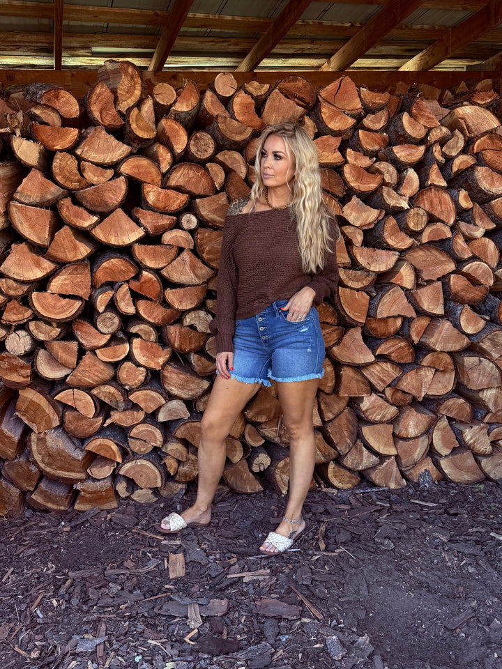 lady wearing chocolate colored open weave off the shoulder sweater with blue jean button fly shorts.