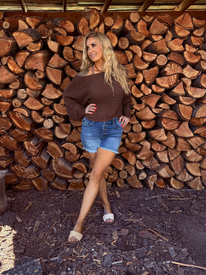 lady wearing chocolate colored open weave off the shoulder sweater with blue jean button fly shorts.