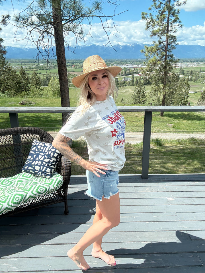 Woman wearing cream colored tshirt that states 'United States - made in america set July 1776 with a cowgirl on her horse' blue and red lettering, denim shorts. 
