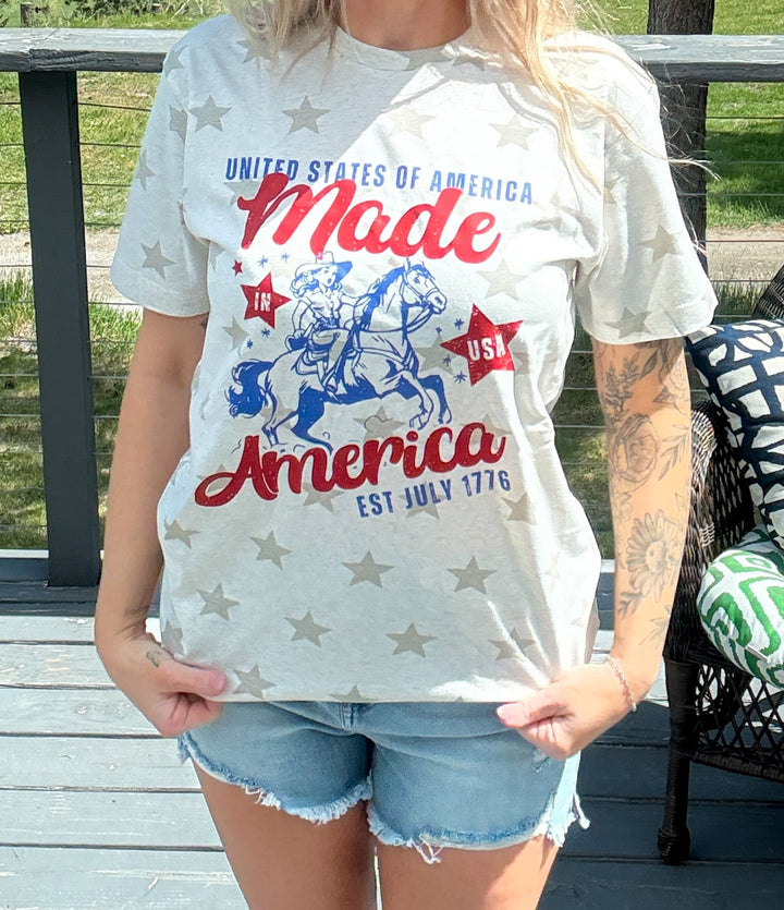 Woman wearing cream colored tshirt that states 'United States - made in america set July 1776 with a cowgirl on her horse' blue and red lettering, denim shorts. 