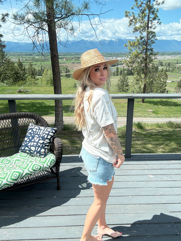 Woman wearing cream colored tshirt that states 'United States - made in america set July 1776 with a cowgirl on her horse' blue and red lettering, denim shorts. 
