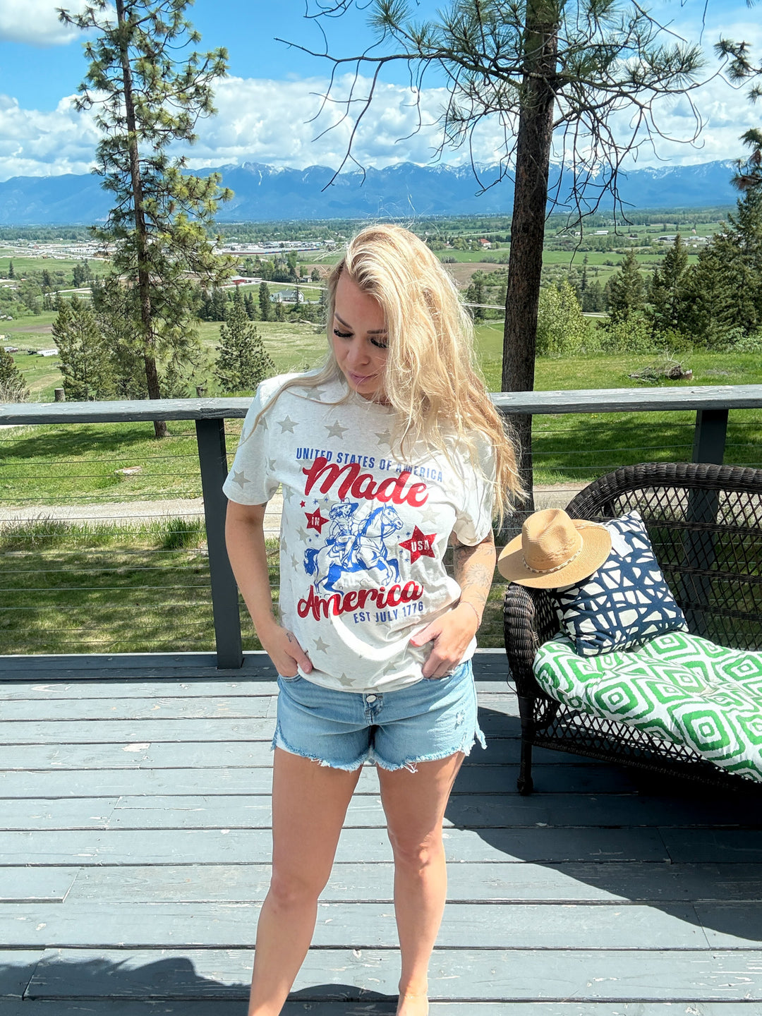 Woman wearing cream colored tshirt that states 'United States - made in america set July 1776 with a cowgirl on her horse' blue and red lettering, denim shorts. 