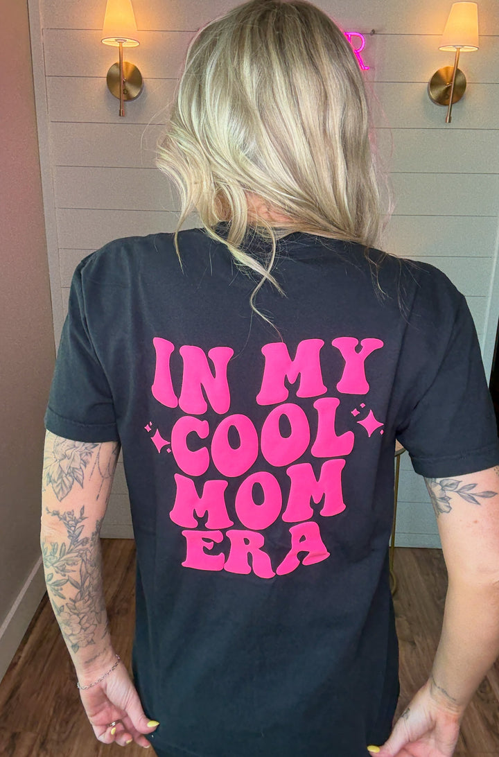 Woman wearing charcoal tee shirt with 'In my cool mom era' in hot pink puff letter graphics and a small hot pink smiley face on the front 