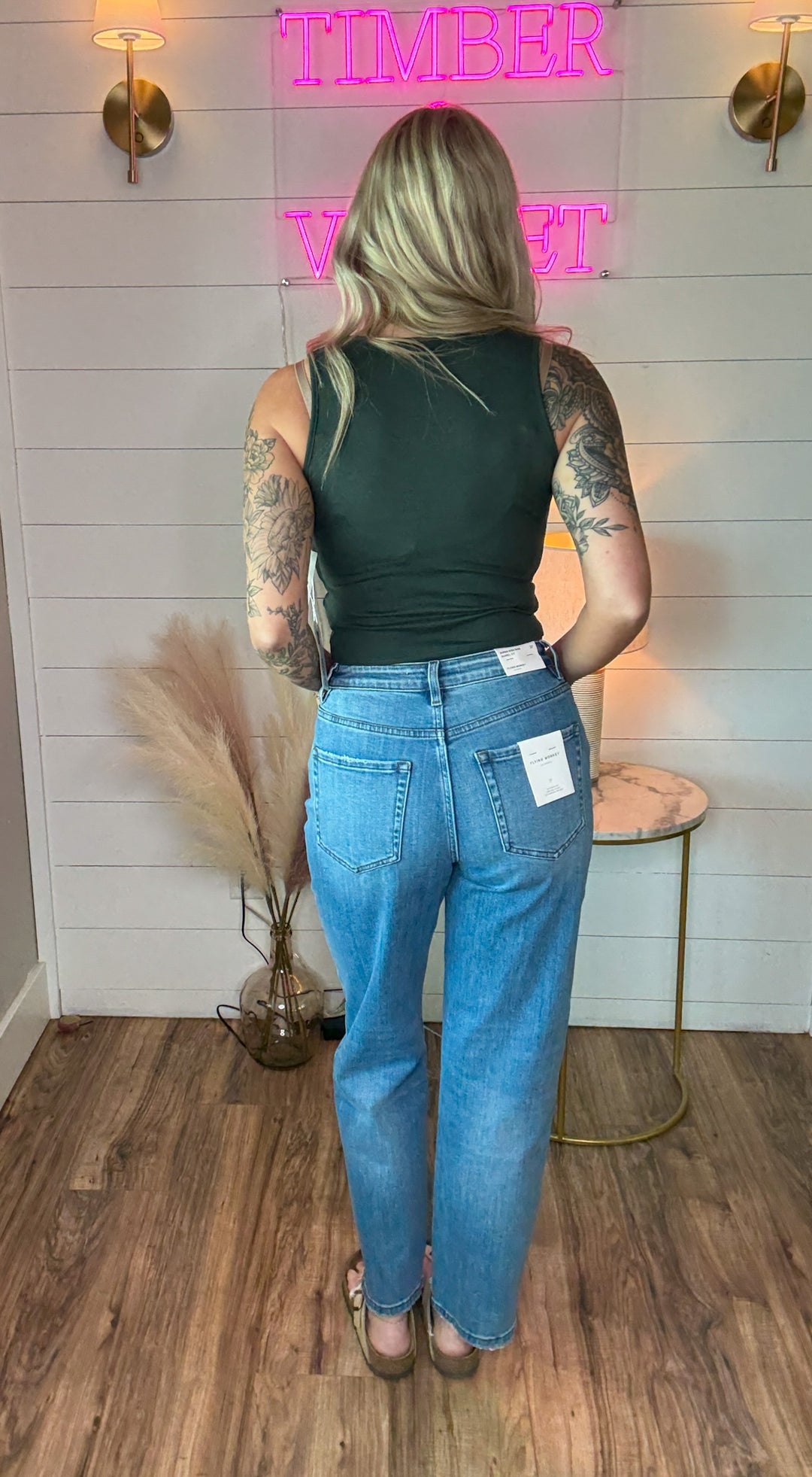 woman wearing a pair of flying monkey flattering barrel cut jeans, light-medium wash, with just the right touch of distressing and raw hem detail. Paired with a green tank top.