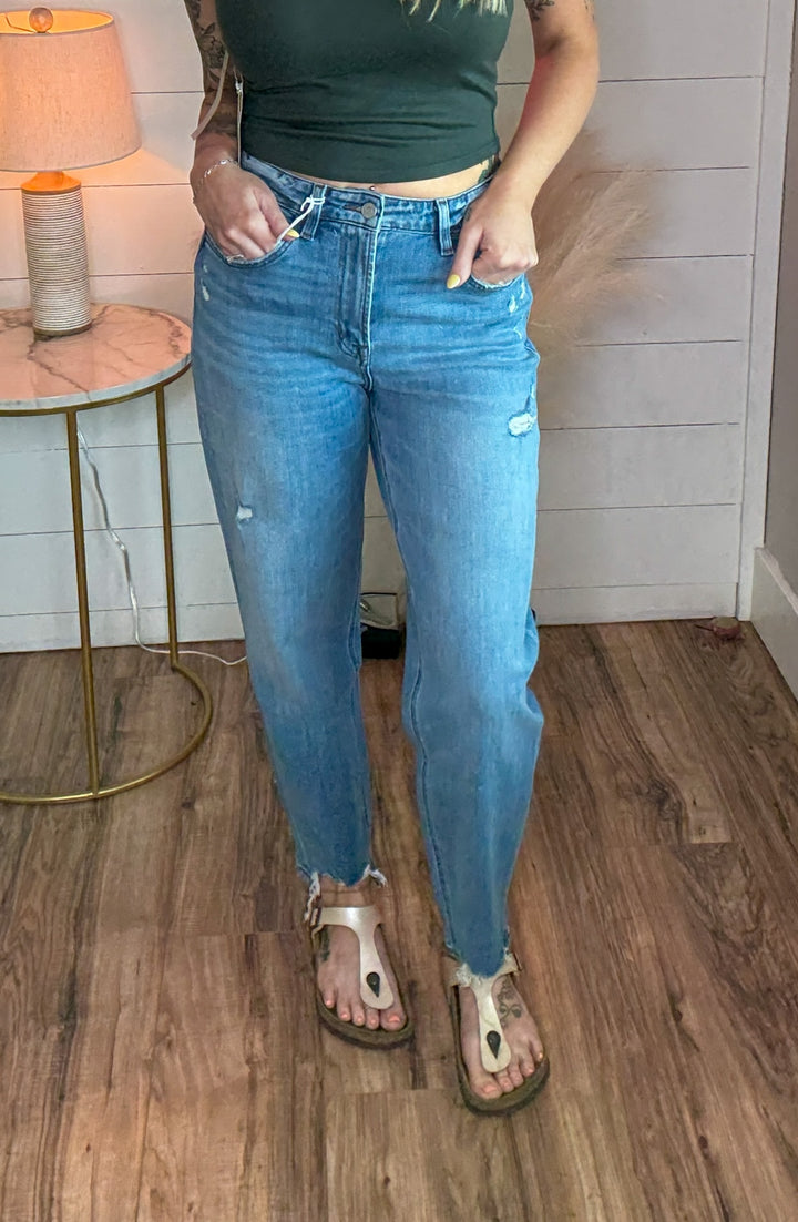 woman wearing a pair of flying monkey flattering barrel cut jeans, light-medium wash, with just the right touch of distressing and raw hem detail. Paired with a green tank top.