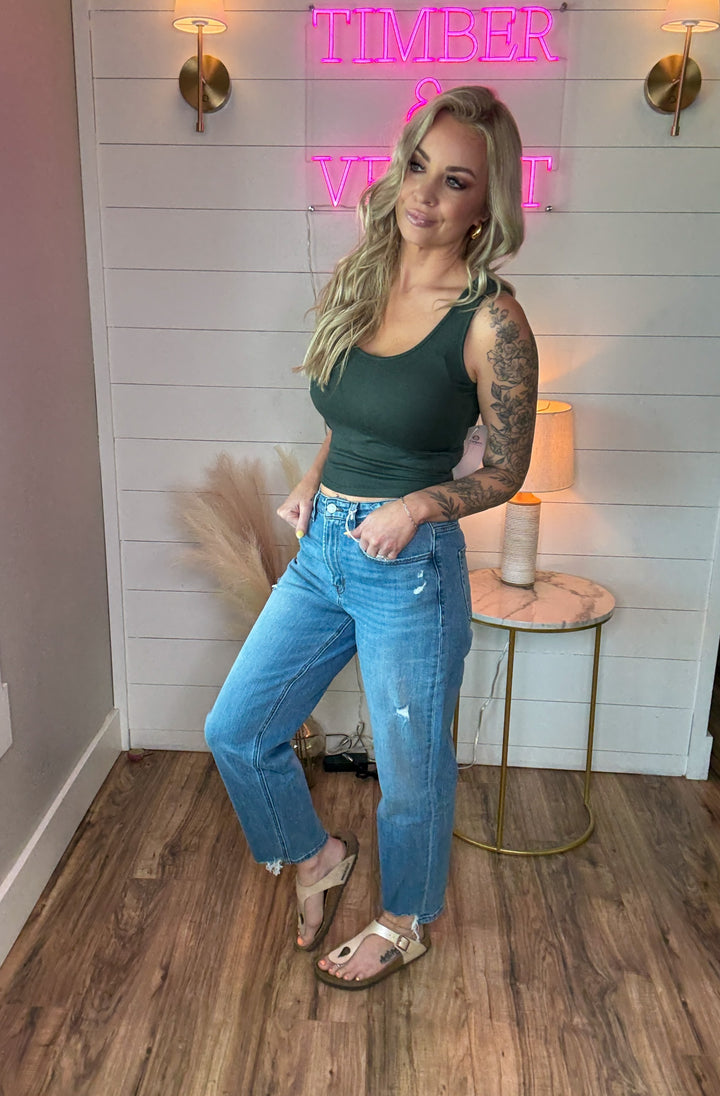 woman wearing a pair of flying monkey flattering barrel cut jeans, light-medium wash, with just the right touch of distressing and raw hem detail. Paired with a green tank top.