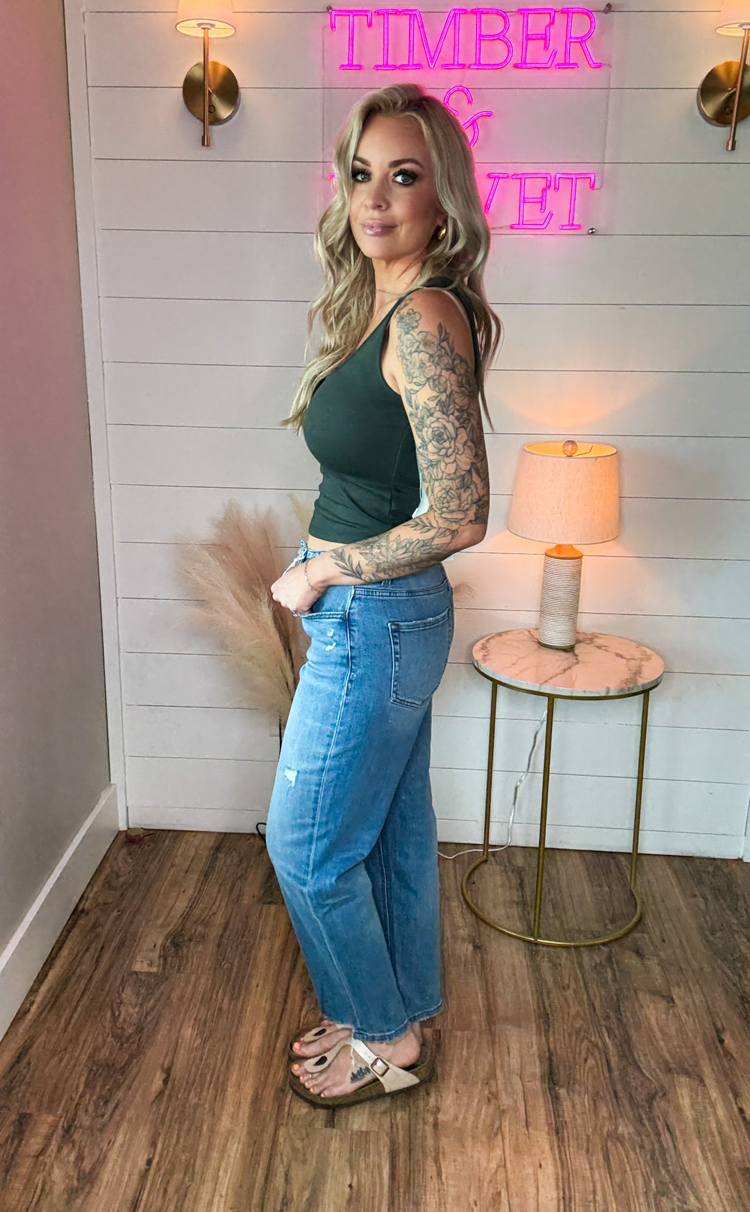 woman wearing a pair of flying monkey flattering barrel cut jeans, light-medium wash, with just the right touch of distressing and raw hem detail. Paired with a green tank top.