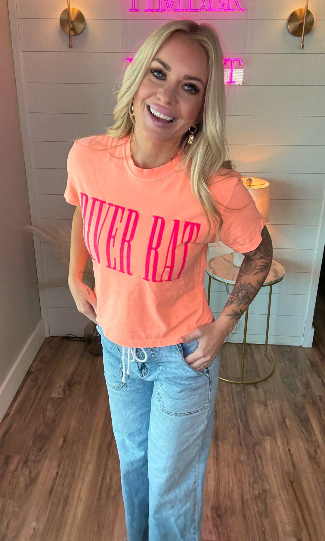 Woman wearing cantaloupe colored slightly cropped tee that says RIVER RAT in pink embossing on the front. Also wearing a pair of blue jeans.