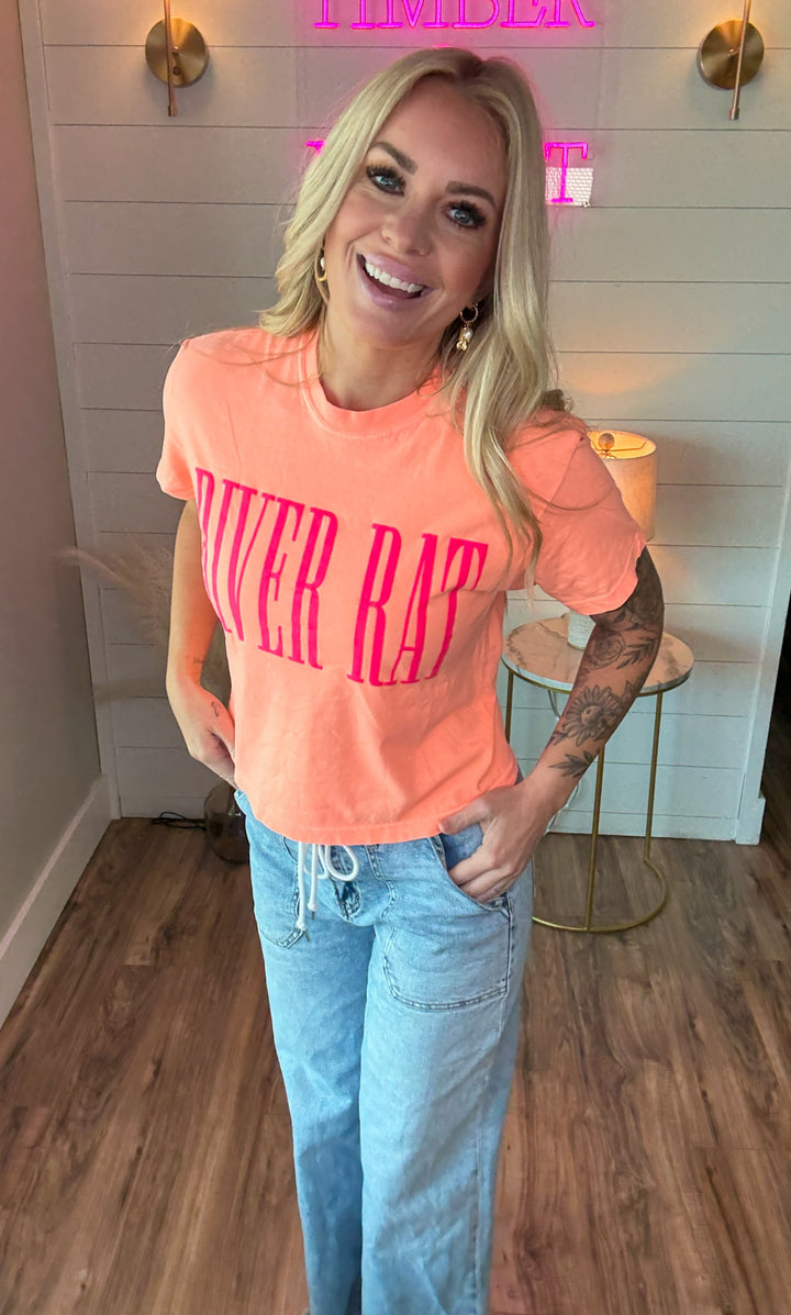 Woman wearing cantaloupe colored slightly cropped tee that says RIVER RAT in pink embossing on the front. Also wearing a pair of blue jeans.