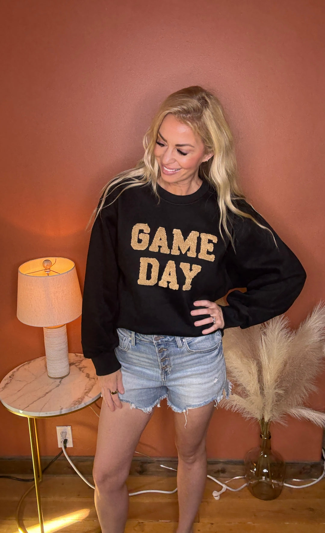 Woman wearing a black sweatshirt with 'GAME DAY' in gold letters, standing in a room with a lamp and decorative items.
