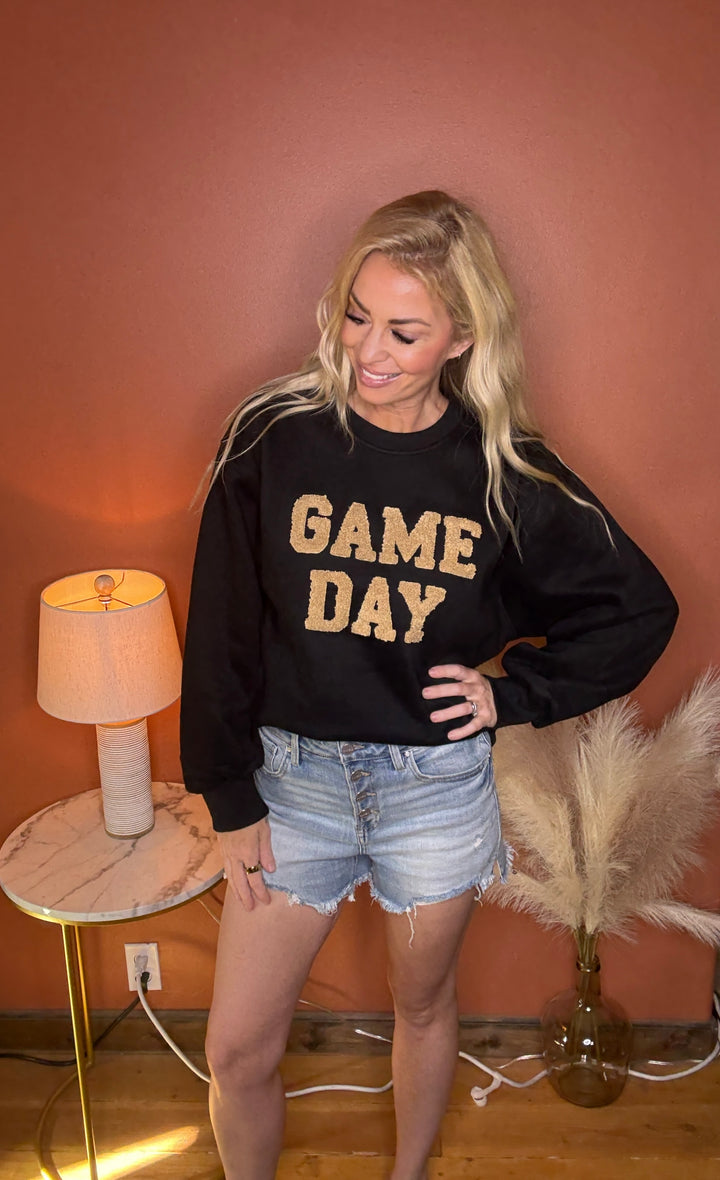 Woman wearing a black sweatshirt with 'GAME DAY' in gold letters, standing in a room with a lamp and decorative items.