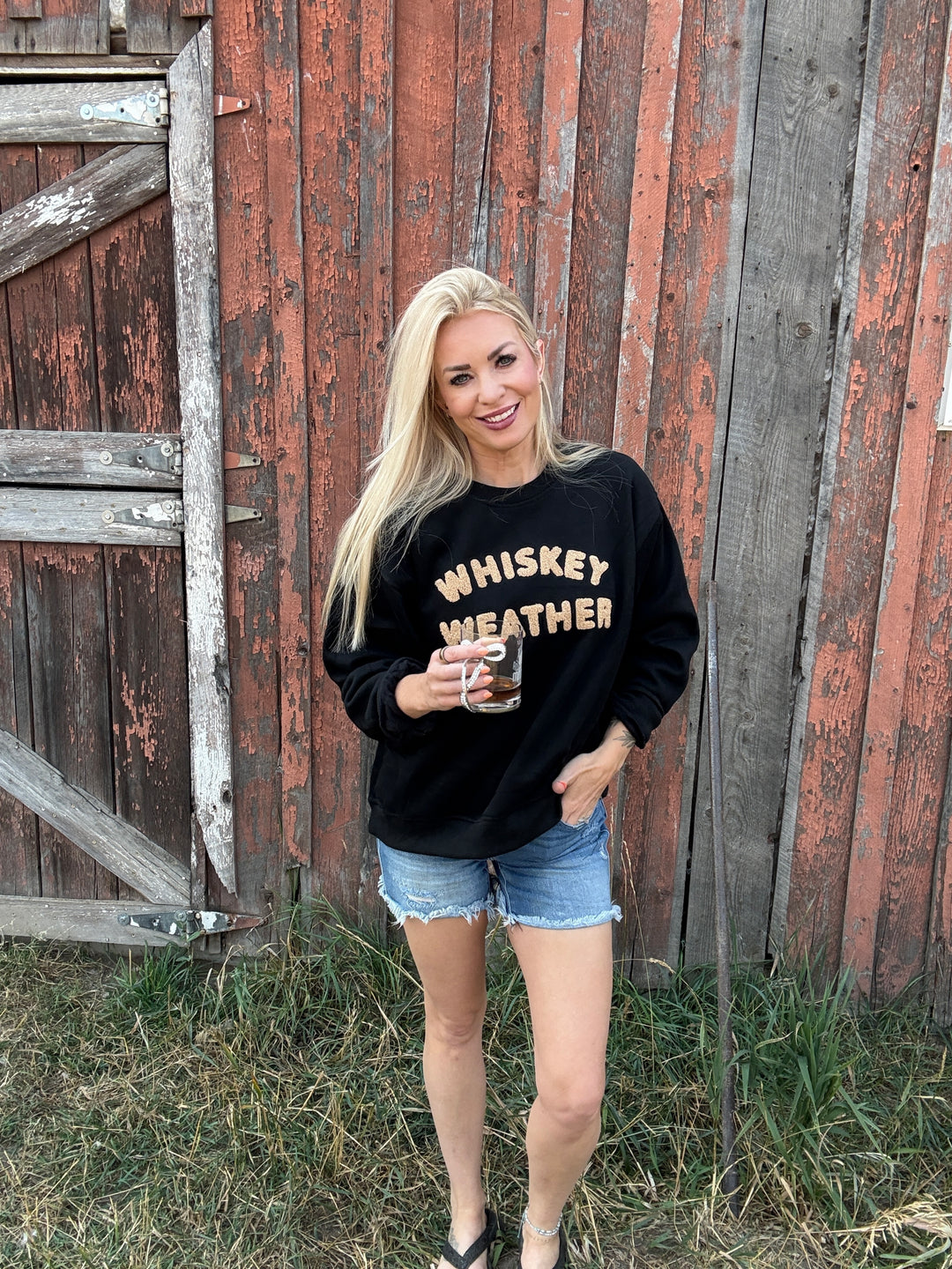 Woman wearing a black sweatshirt with 'WHISKEY WEATHER' text, standing in front of a rustic wooden building.