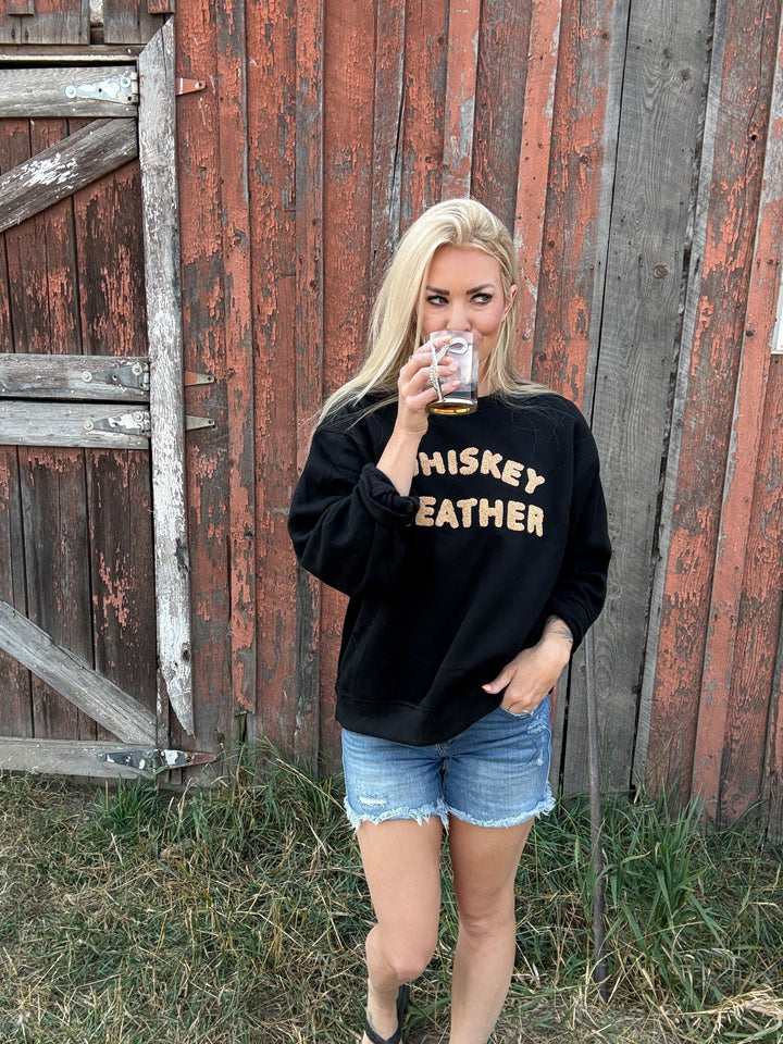 Person wearing a black sweatshirt with 'Whiskey Weather' text, standing in front of a rustic wooden wall.