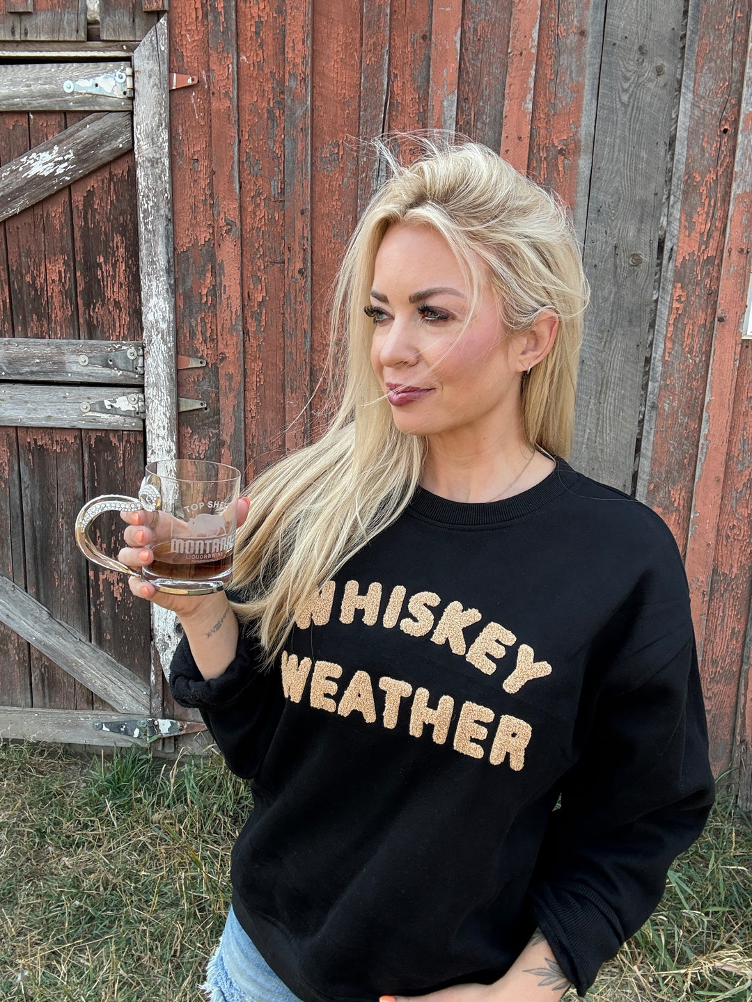 Woman wearing a black sweatshirt with 'WHISKEY WEATHER' text, holding a glass of whiskey in front of a wooden fence.