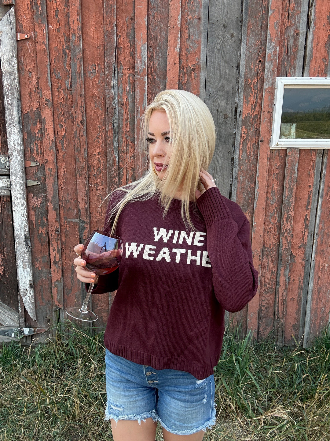 Person wearing a maroon sweater with 'WINE WEATHER' text, holding a glass of wine, standing in front of a rustic wooden building.