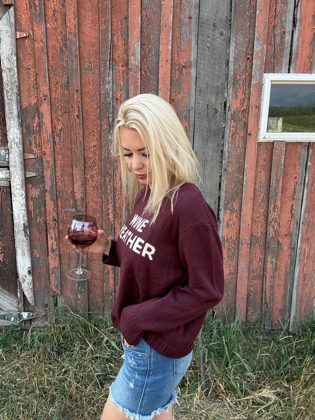 Woman in maroon sweatshirt wearing a 'Wine Weather' sweater holding a glass of red wine in front of a rustic wooden building.
