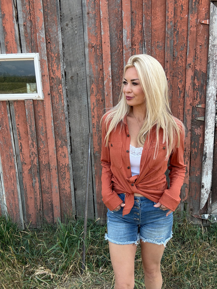 Woman standing in front of a rustic wooden building wearing an rust colored button up and denim shorts.