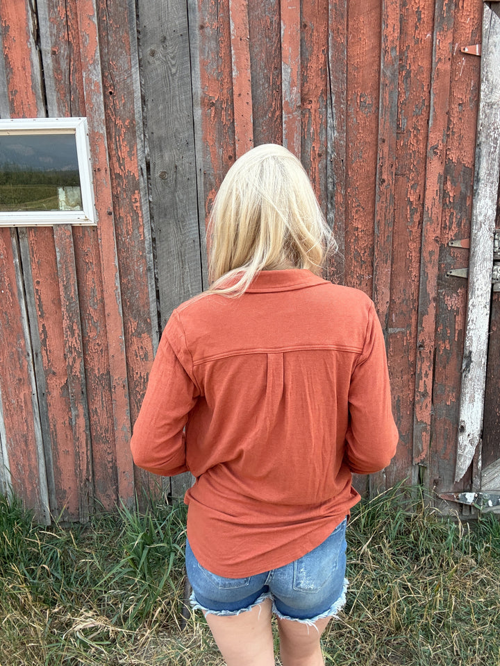Person wearing a rust colored button up shirt and denim shorts standing in front of a rustic wooden building.
