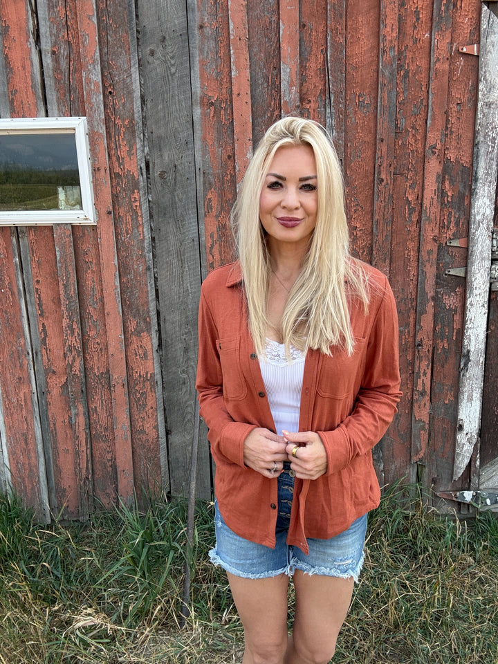 Woman standing in front of a rustic wooden building wearing a rust colored button up and denim shorts.