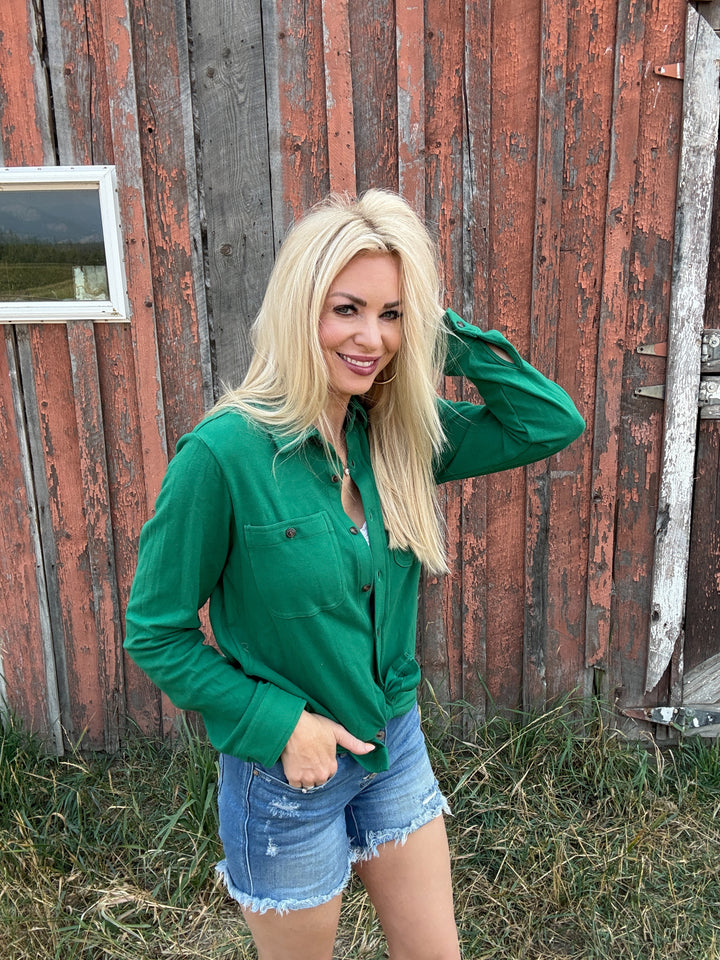 Woman in a green button up shirt and denim shorts standing in front of a rustic wooden building.