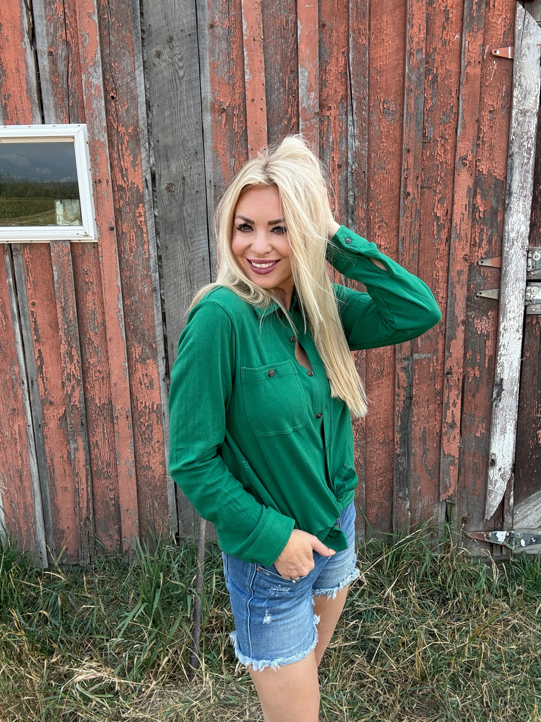 Woman in a green button up shirt and denim shorts standing in front of a rustic wooden building.