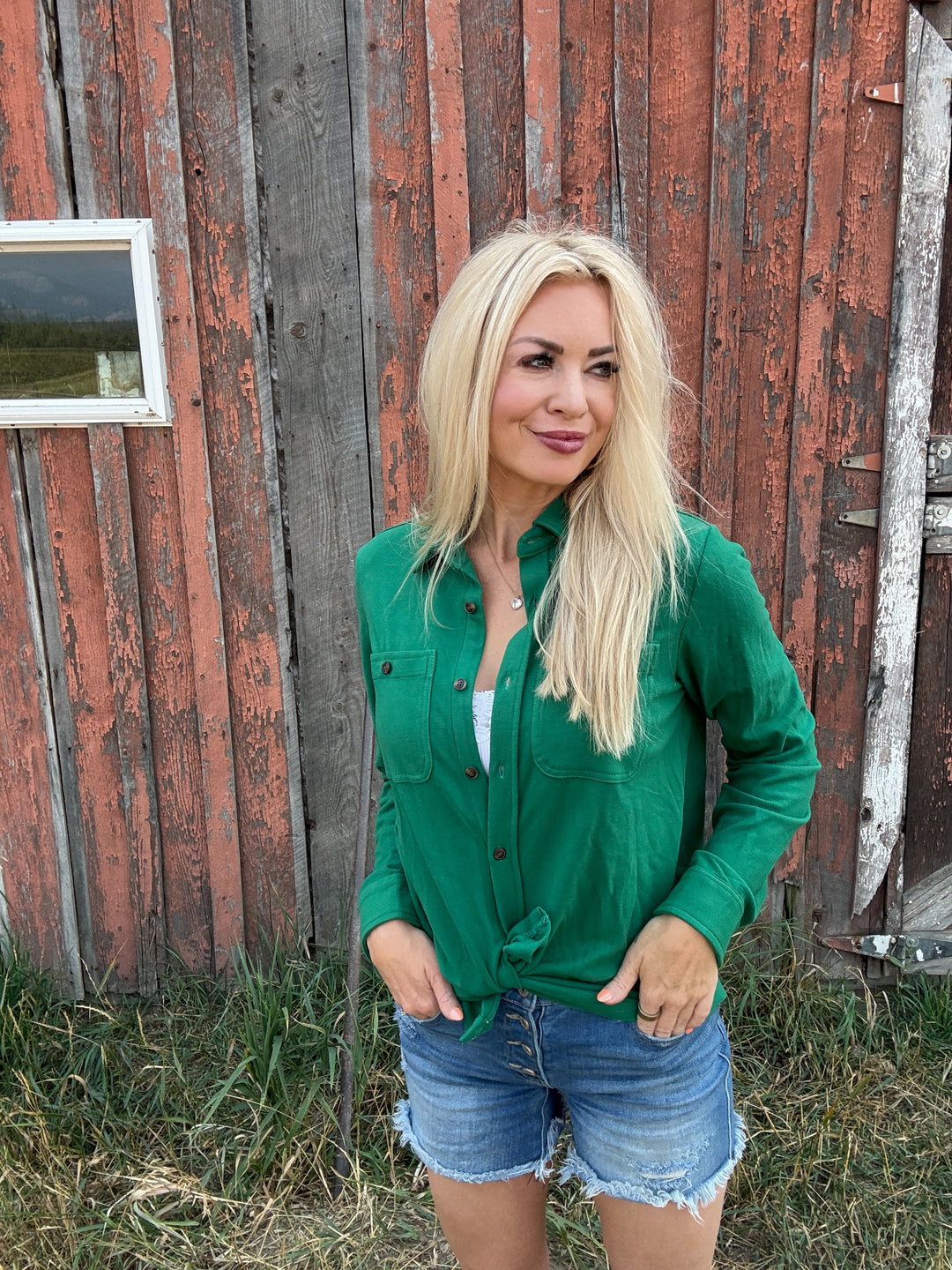 Woman in a green button up shirt and denim shorts standing in front of a rustic wooden building.