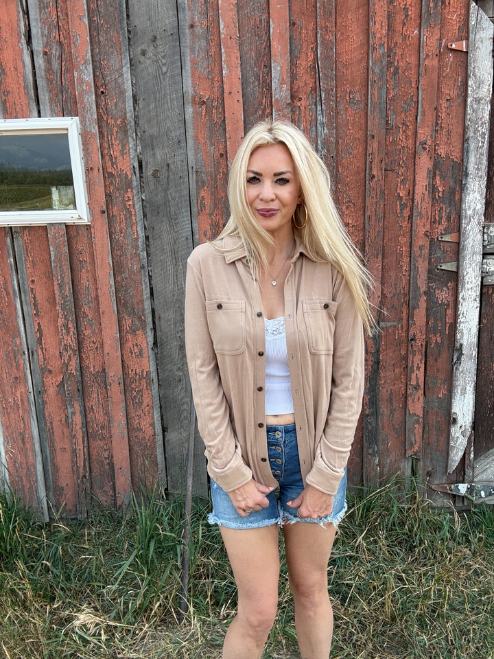 Woman standing in front of a rustic wooden building wearing a taupe button up and denim shorts.
