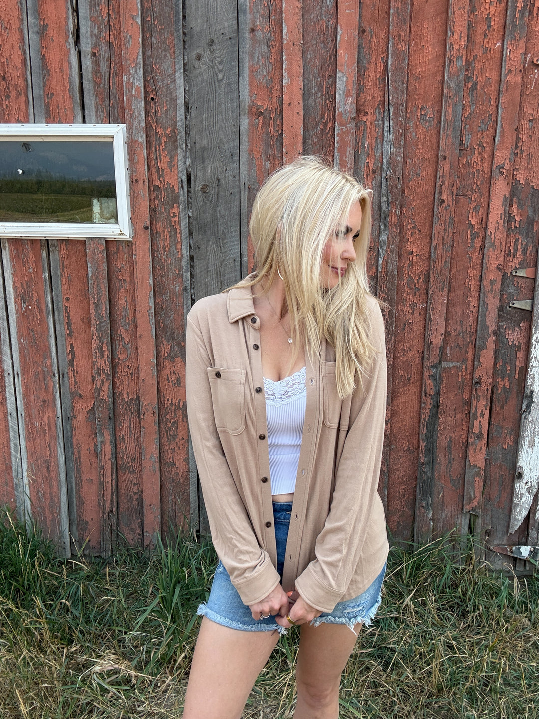 Woman standing in front of a rustic red barn wearing a taupe button up shirt and denim shorts 