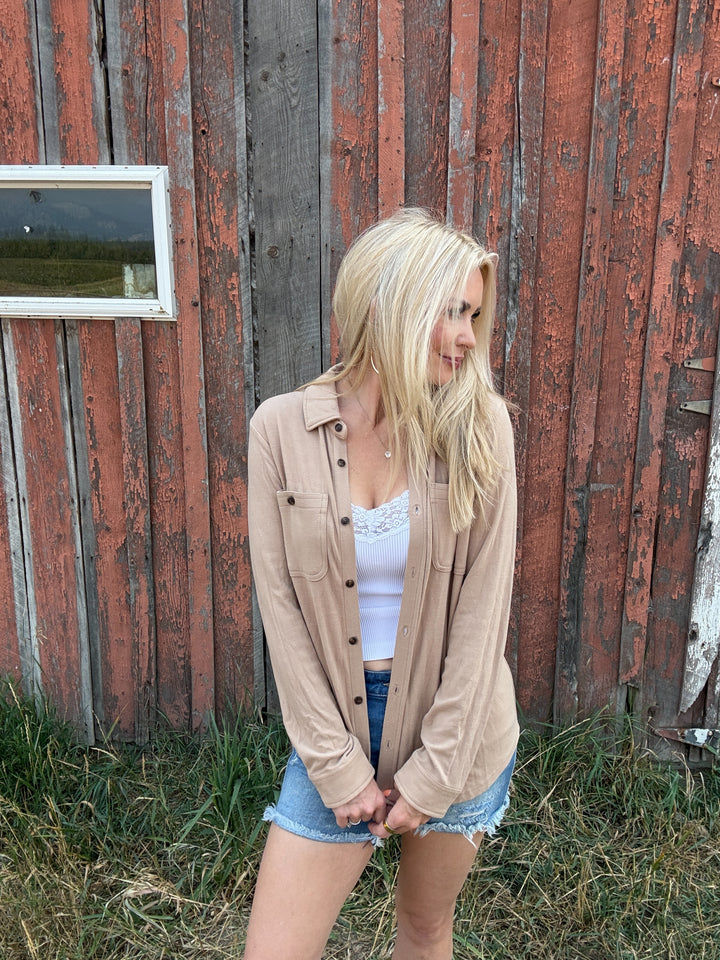 Woman standing in front of a rustic red barn wearing a taupe button up shirt and denim shorts 