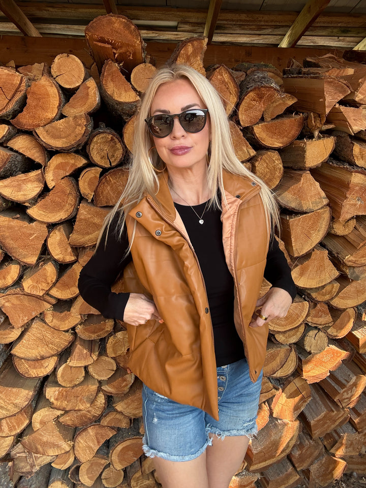 Woman wearing a camel brown faux leather vest and sunglasses standing in front of stacked logs.