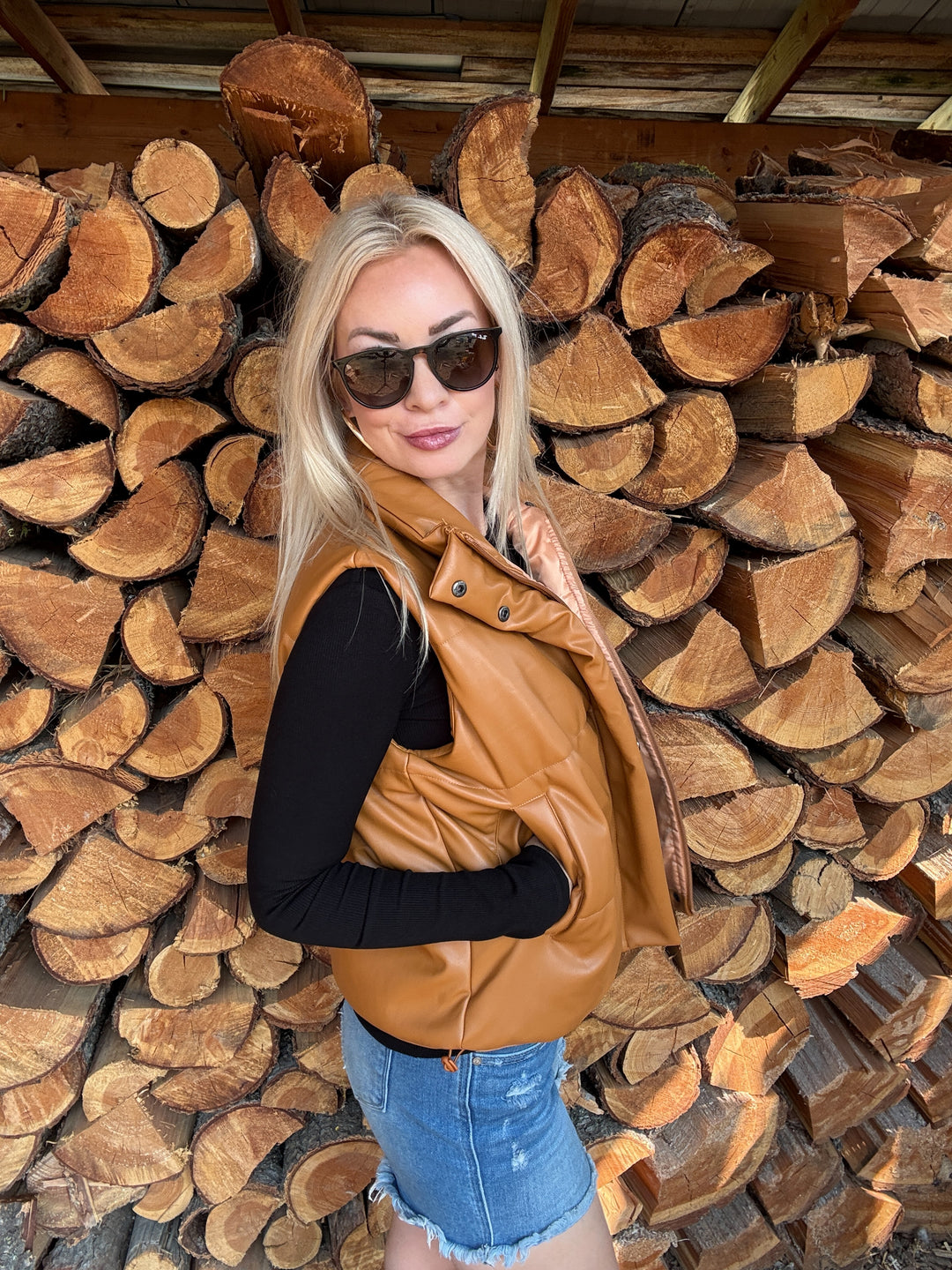 Woman wearing a camel brown faux leather vest and sunglasses standing in front of stacked logs.