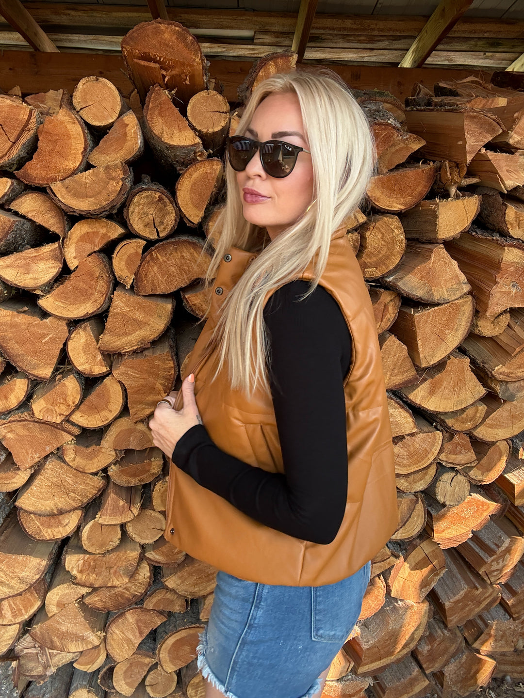 Woman wearing a camel brown faux leather vest and sunglasses standing in front of stacked logs.