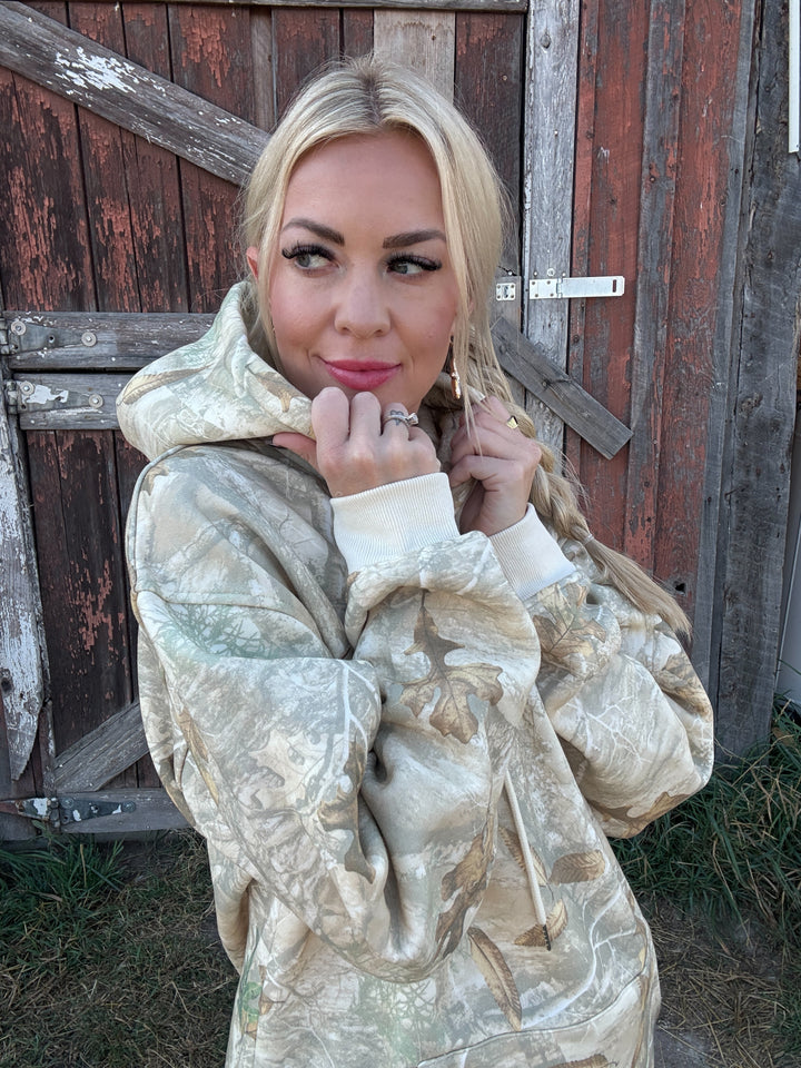 Woman wearing a camo MONTANA hoodie in front of a wooden door.