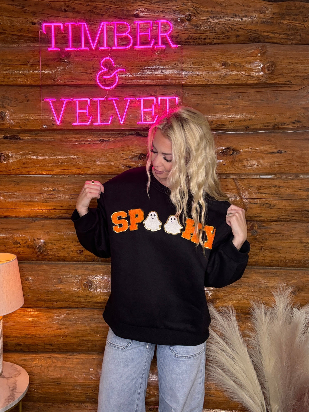 Person wearing a black sweatshirt with colorful text that says spooky in front of a wooden wall with 'Timber & Velvet' neon sign.